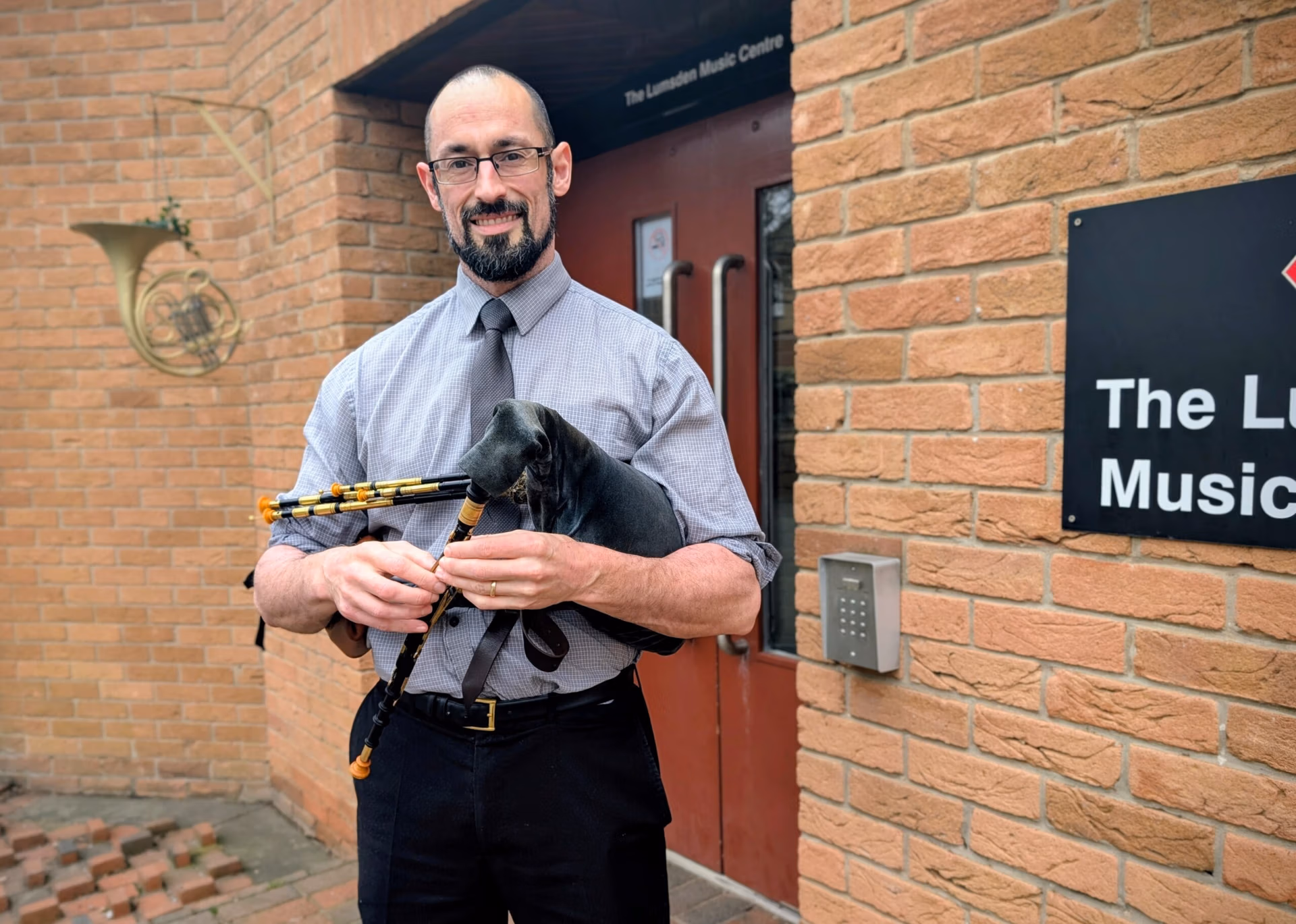 The image shows Dame Allan's Schools Head of Latin, Andy Lawrenson, in front of a brick building holding Northumbrian smallpipes (like small bagpipes). He is in a blue shirt, tall and bearded with dark hair and glasses.